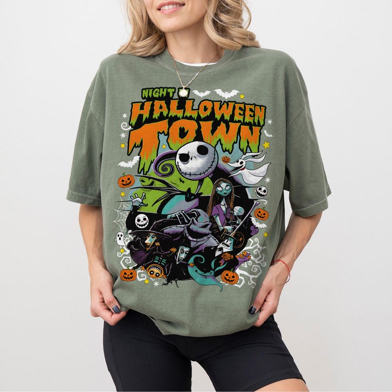 May include: Olive green t-shirt with a colorful graphic featuring characters from "The Nightmare Before Christmas." The design includes Jack Skellington, Sally, and other Halloween Town figures, with the text "Night Halloween Town."