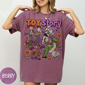 Op de afbeelding: Een bessenkleurig t-shirt met een Toy Story Halloween-afbeelding met de personages Jessie, Woody, Buzz Lightyear, Rex, Hamm, Slinky Dog en Bullseye. Het ontwerp bevat pompoenen, maissnoepjes, vleermuizen en de tekst Toy Story Since 1995.