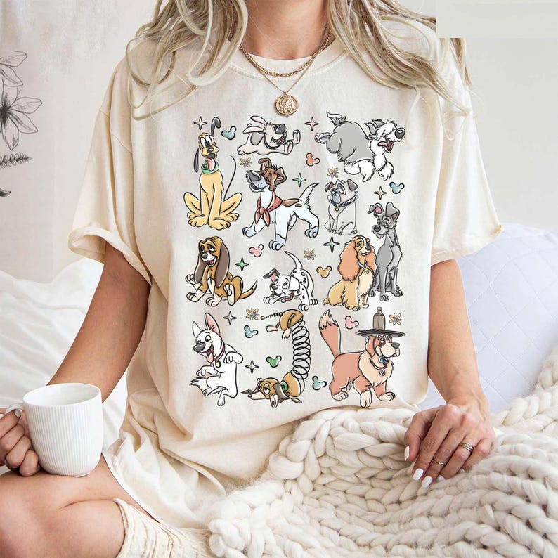 May include: Cream-colored t-shirt featuring a colorful cartoon illustration of various dog breeds. The dogs are depicted in a playful, animated style, with a variety of poses and expressions. The shirt has short sleeves and a relaxed fit.