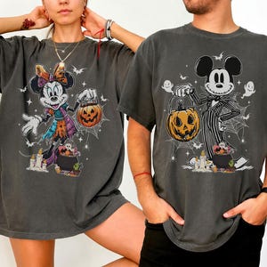 May include: Two dark gray t-shirts featuring Halloween-themed Disney characters. One shirt displays Minnie Mouse in a witch costume holding a jack-o'-lantern. The other shirt shows Mickey Mouse as Jack Skellington, also holding a pumpkin.