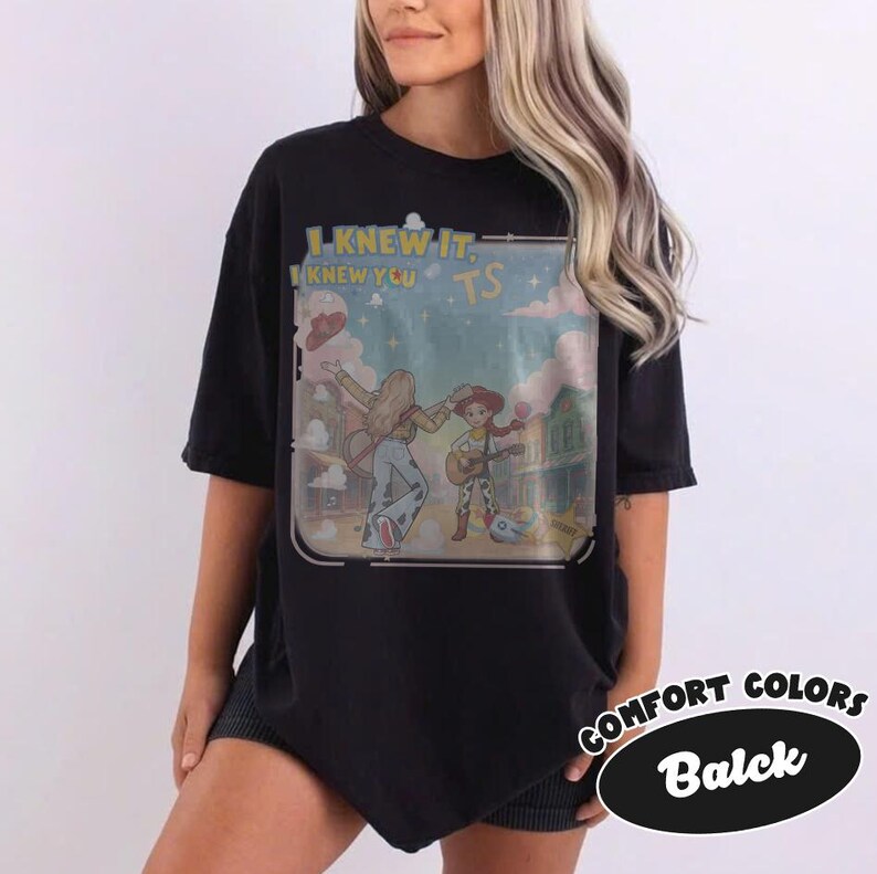 May include: A black t-shirt featuring a graphic print of two figures in a western-style setting with guitars. The text "I KNEW IT. I KNEW YOU TS" is printed in yellow above the illustration, which includes a blue sky, clouds, and buildings.
