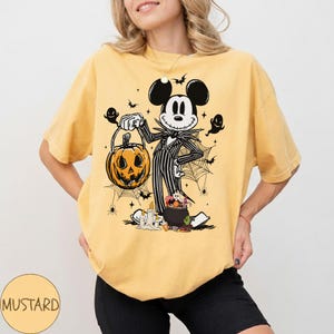 May include: Mustard-colored t-shirt featuring a Halloween-themed graphic. The graphic depicts Mickey Mouse dressed as Jack Skellington, holding a carved pumpkin. Spiders, bats, and a cauldron with candy complete the design. The word "MUSTARD" is printed on the bottom left.