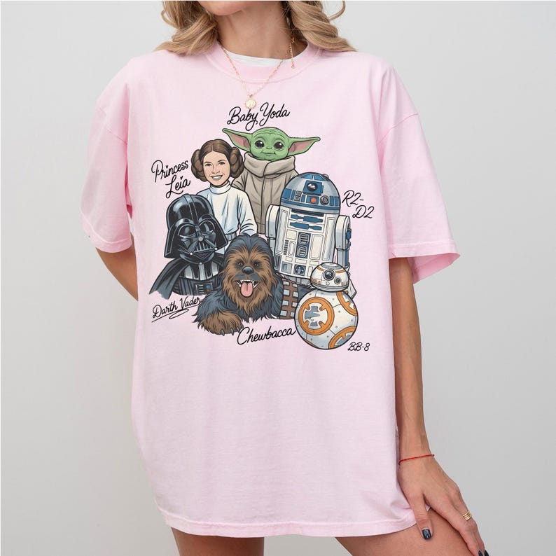 May include: A pale pink t-shirt with a graphic of Star Wars characters, including Darth Vader, Princess Leia, Baby Yoda, Chewbacca, R2-D2, and BB-8. The names of the characters are written in a stylized font. The shirt has short sleeves and a crew neck.