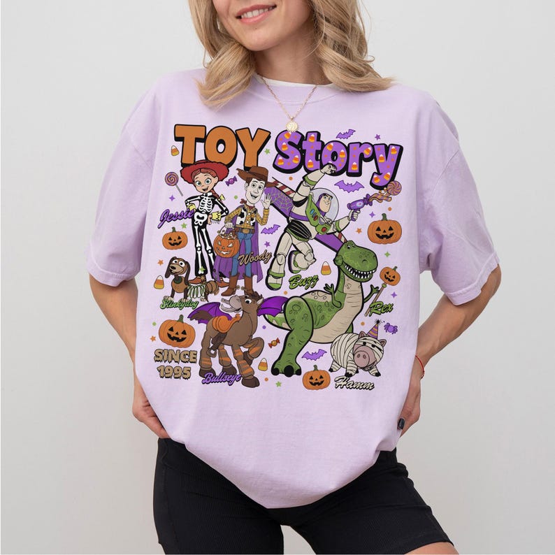 Op de afbeelding: Een lichtpaars t-shirt met een Toy Story Halloween-afbeelding met de personages Jessie, Woody, Buzz Lightyear, Slinky Dog, Bullseye, Rex en Hamm. Het ontwerp bevat pompoenen, maissnoepjes, vleermuizen en de tekst Toy Story Since 1995.