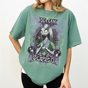 K&ouml;nnte beinhalten: Salbeigr&uuml;nes T-Shirt mit einer Grafik einer Figur mit langen wei&szlig;en Haaren, gen&auml;hten Merkmalen und einem gr&uuml;nen Kleid. Der Text "SCREAM QUEEN" wird oberhalb und unterhalb der Figur in einem lila-wei&szlig;en Design angezeigt.