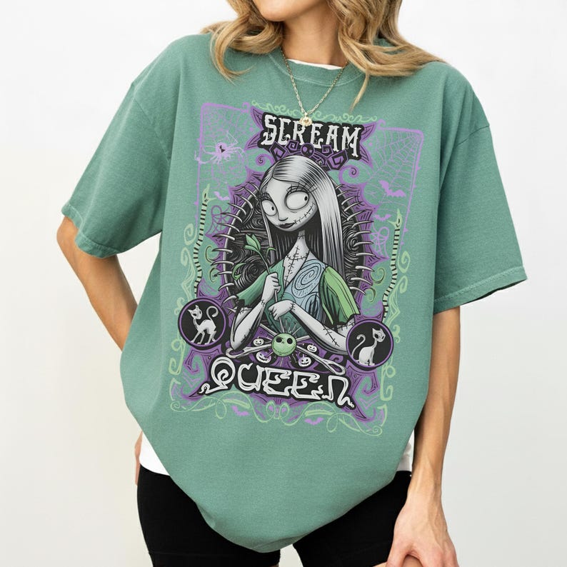 K&ouml;nnte beinhalten: Salbeigr&uuml;nes T-Shirt mit einer Grafik einer Figur mit langen wei&szlig;en Haaren, gen&auml;hten Merkmalen und einem gr&uuml;nen Kleid. Der Text "SCREAM QUEEN" wird oberhalb und unterhalb der Figur in einem lila-wei&szlig;en Design angezeigt.