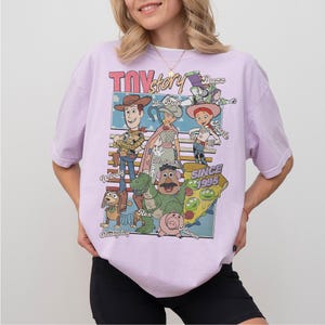 Pu&ograve; includere: Una maglietta lilla con una grafica dei personaggi del film d'animazione "Toy Story". Il design include Woody, Buzz Lightyear, Jessie e altri personaggi, con il testo "TOY STORY" e "SINCE 1995."