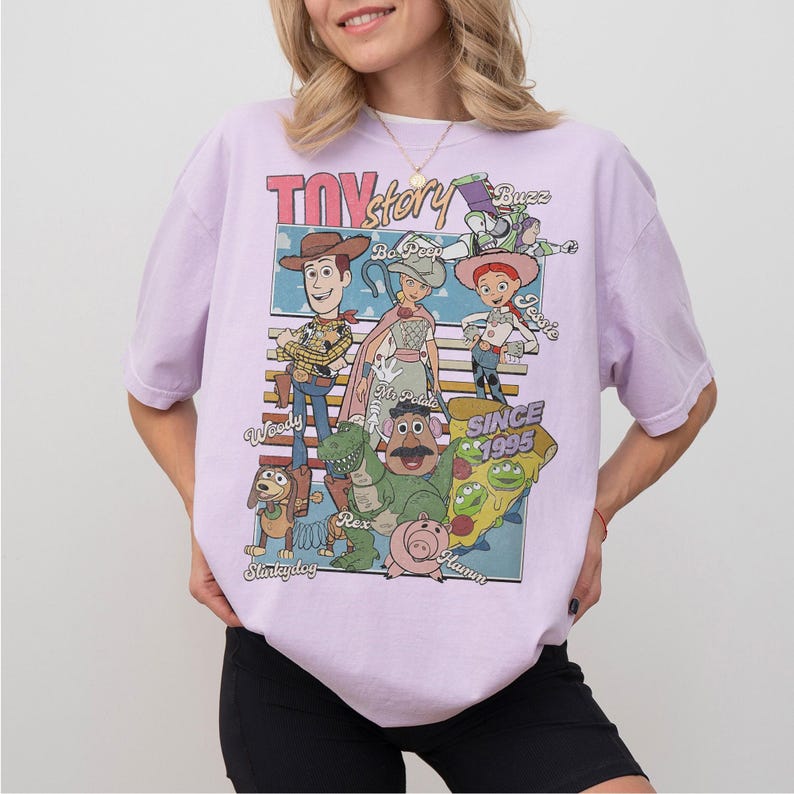 Pu&ograve; includere: Una maglietta lilla con una grafica dei personaggi del film d'animazione "Toy Story". Il design include Woody, Buzz Lightyear, Jessie e altri personaggi, con il testo "TOY STORY" e "SINCE 1995."