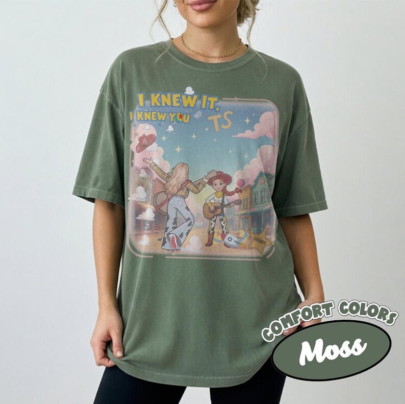 May include: A moss green short-sleeve t-shirt featuring a graphic print of two cartoon characters playing guitars in a western town setting. The text "I KNEW IT, I KNEW YOU TS" is printed above the characters against a pastel sky with clouds.