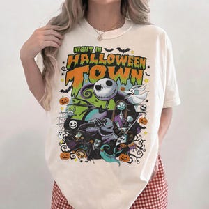 May include: Cream-colored t-shirt featuring a colorful graphic from "The Nightmare Before Christmas." The design includes Jack Skellington, Sally, and other characters with the text "Night in Halloween Town."