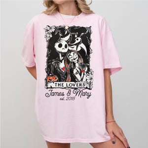 May include: A light pink t-shirt featuring a black and white graphic of Jack and Sally from The Nightmare Before Christmas. The graphic includes the text "THE LOVERS" and "James & Mary est. 2018". The shirt has short sleeves.