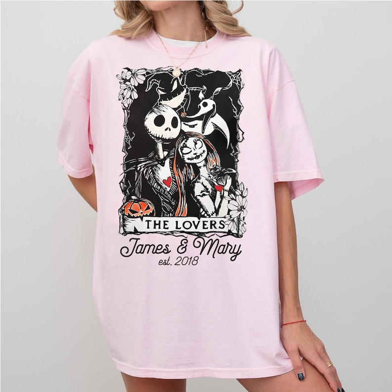 May include: A light pink t-shirt featuring a black and white graphic of Jack and Sally from The Nightmare Before Christmas. The graphic includes the text "THE LOVERS" and "James & Mary est. 2018". The shirt has short sleeves.