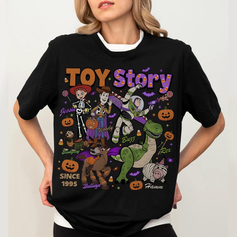 Op de afbeelding: Een zwart t-shirt met een Toy Story Halloween-afbeelding. Het ontwerp bevat de personages Jessie, Woody, Buzz Lightyear, Rex, Bullseye, Slinky Dog en Hamm, omringd door pompoenen, snoepma&iuml;s en vleermuizen. De tekst luidt Toy Story en Since 1995.