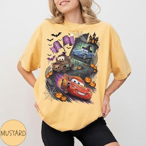 K&ouml;nnte beinhalten: Ein senfgelbes Kurzarm-T-Shirt mit einem Grafikdruck von zwei Cartoon-Autos auf einer kurvenreichen Stra&szlig;e. Das Design zeigt einen braunen Abschleppwagen, ein rotes Rennauto, K&uuml;rbisse, Spinnennetze, Flederm&auml;use und die Silhouette eines Spukhauses.
