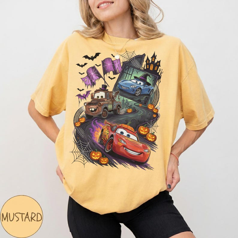 K&ouml;nnte beinhalten: Ein senfgelbes Kurzarm-T-Shirt mit einem Grafikdruck von zwei Cartoon-Autos auf einer kurvenreichen Stra&szlig;e. Das Design zeigt einen braunen Abschleppwagen, ein rotes Rennauto, K&uuml;rbisse, Spinnennetze, Flederm&auml;use und die Silhouette eines Spukhauses.