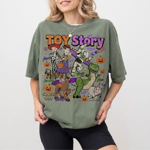 Op de afbeelding: Een olijfgroen t-shirt met een grafische print van Toy Story-personages in Halloween-kostuums. Het ontwerp bevat Woody, Buzz Lightyear, Jessie, Rex, Bullseye, Slinky Dog en Hamm, omringd door pompoenen, maissnoepjes en vleermuizen.