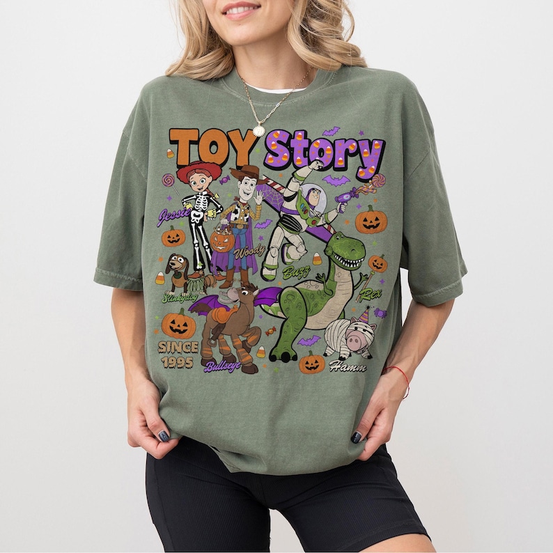 Op de afbeelding: Een olijfgroen t-shirt met een grafische print van Toy Story-personages in Halloween-kostuums. Het ontwerp bevat Woody, Buzz Lightyear, Jessie, Rex, Bullseye, Slinky Dog en Hamm, omringd door pompoenen, maissnoepjes en vleermuizen.