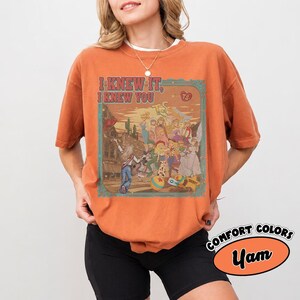 Op de afbeelding: Een roestoranje t-shirt met korte mouwen en een grafische print met de tekst I KNEW IT, I KNEW YOU. De illustratie toont een groep geanimeerde personages in een woestijnomgeving met een gebouw, cactussen en een stervormig badge-icoon.