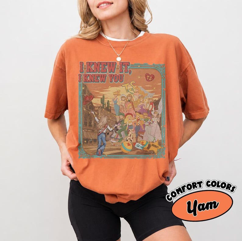 Op de afbeelding: Een roestoranje t-shirt met korte mouwen en een grafische print met de tekst I KNEW IT, I KNEW YOU. De illustratie toont een groep geanimeerde personages in een woestijnomgeving met een gebouw, cactussen en een stervormig badge-icoon.