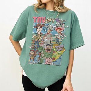 Pu&ograve; includere: T-shirt verde con grafica di Toy Story con Woody, Buzz e Jessie. Il design include il testo "Toy Story" e "Since 1995". La maglietta ha maniche corte e girocollo.