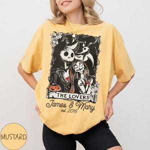 May include: Mustard-colored t-shirt featuring a black and white graphic of Jack Skellington and Sally from The Nightmare Before Christmas. The graphic includes the text "THE LOVERS" and "James & Mary est. 2018".