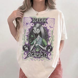 K&ouml;nnte beinhalten: Cremefarbenes T-Shirt mit einer Grafik einer Figur mit langen schwarzen Haaren und einem gr&uuml;nen Kleid. Das Design enth&auml;lt den Text "SCREAM QUEEN" in einem lila-gr&uuml;nen gotischen Stil mit Spinnweben und Katzensilhouetten.