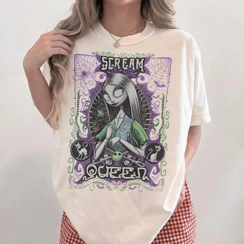 K&ouml;nnte beinhalten: Cremefarbenes T-Shirt mit einer Grafik einer Figur mit langen schwarzen Haaren und einem gr&uuml;nen Kleid. Das Design enth&auml;lt den Text "SCREAM QUEEN" in einem lila-gr&uuml;nen gotischen Stil mit Spinnweben und Katzensilhouetten.