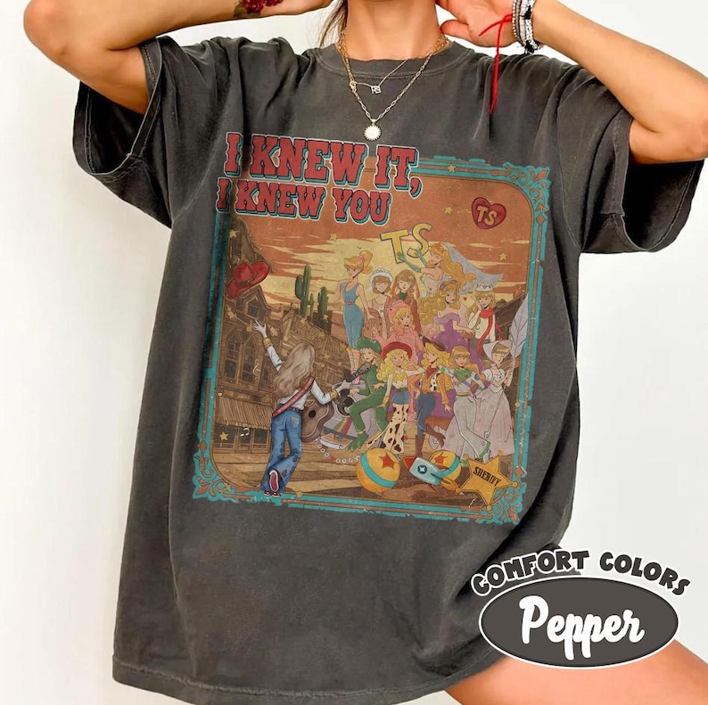Op de afbeelding: Een antracietgrijs t-shirt met korte mouwen en een grafische print met de tekst I KNEW IT, I KNEW YOU. De illustratie toont een groep geanimeerde figuren in een woestijnomgeving met een gitaar, een ster en een hart-icoon met de letters TS.