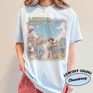 May include: A light blue t-shirt featuring a graphic print of two cartoon characters playing guitars in a western town setting. The text "I KNEW IT, I KNEW YOU" and "TS" is printed above the characters. The design includes pink clouds and stars.