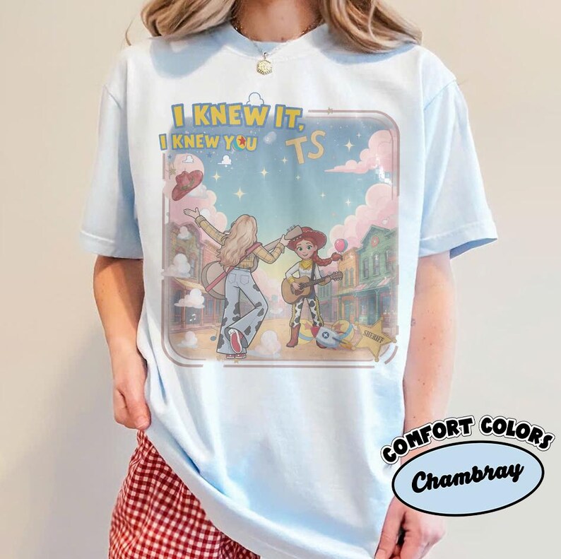 May include: A light blue t-shirt featuring a graphic print of two cartoon characters playing guitars in a western town setting. The text "I KNEW IT, I KNEW YOU" and "TS" is printed above the characters. The design includes pink clouds and stars.