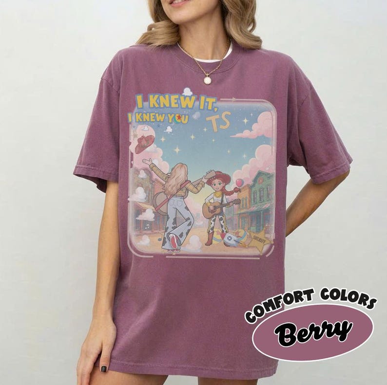 May include: A berry-colored short-sleeve t-shirt featuring a graphic print of two cartoon characters playing guitars in a western town setting. The text "I KNEW IT, I KNEW YOU TS" is printed above the characters. The shirt has a crew neckline.