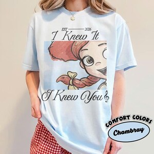 May include: A light blue t-shirt featuring a graphic of a cartoon character with red hair and a braid. The text reads "EST 2026", "I Knew It", and "I Knew You" with a musical note symbol. The shirt has a crew neckline and short sleeves.