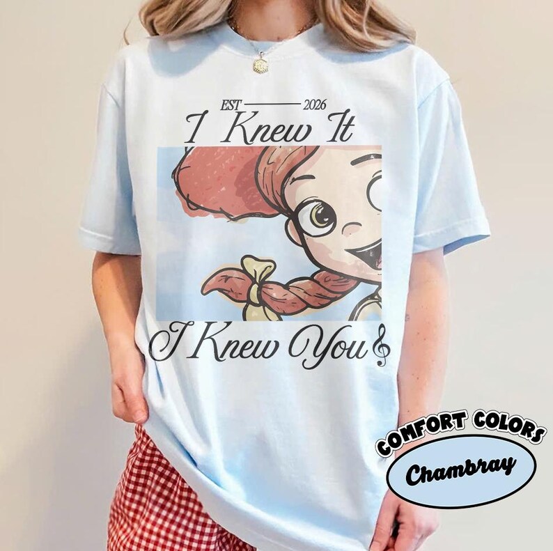 May include: A light blue t-shirt featuring a graphic of a cartoon character with red hair and a braid. The text reads "EST 2026", "I Knew It", and "I Knew You" with a musical note symbol. The shirt has a crew neckline and short sleeves.