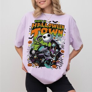 May include: A light purple t-shirt featuring a colorful graphic from "The Nightmare Before Christmas." The design includes Jack Skellington, Sally, and other characters, with the text "Night in Halloween Town."