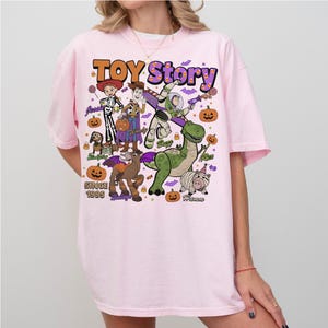 Op de afbeelding: Een lichtroze t-shirt met een kleurrijke Toy Story Halloween-afbeelding. De print bevat de personages Jessie, Woody, Buzz Lightyear, Rex, Slinky Dog, Bullseye en Hamm, omringd door pompoenen, snoepma&iuml;s en vleermuizen met de tekst Toy Story.