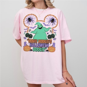 May include: A light pink t-shirt featuring a colorful graphic design. The design includes a green Oogie Boogie character, spiders, bats, dice, and pumpkins. The text "Oogie Boogie's Halloween Boogie" is also displayed.