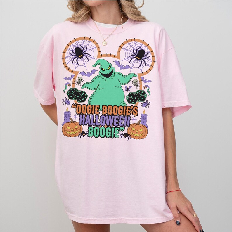 May include: A light pink t-shirt featuring a colorful graphic design. The design includes a green Oogie Boogie character, spiders, bats, dice, and pumpkins. The text "Oogie Boogie's Halloween Boogie" is also displayed.