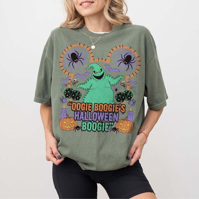 May include: Olive green t-shirt featuring a graphic of Oogie Boogie from The Nightmare Before Christmas. The design includes spiders, dice, pumpkins, and the text "Oogie Boogie's Halloween Boogie".