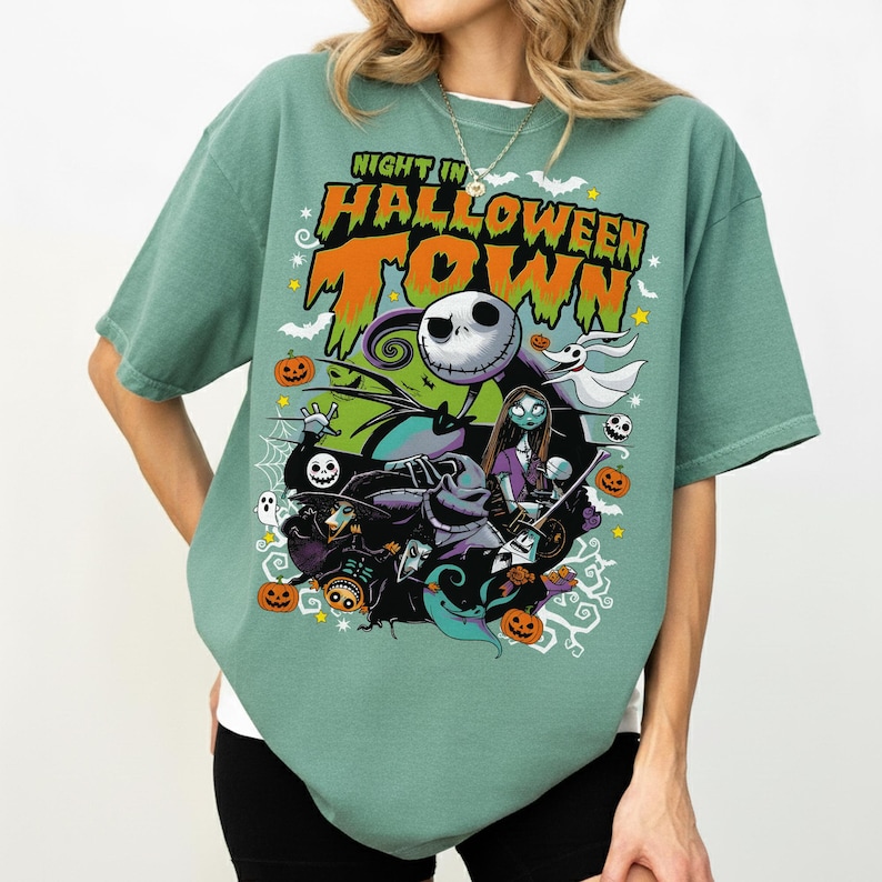 May include: Sage green t-shirt featuring a colorful graphic from "The Nightmare Before Christmas." The design includes Jack Skellington, Sally, and other characters with the text "Night in Halloween Town." Pumpkins and bats are also featured.