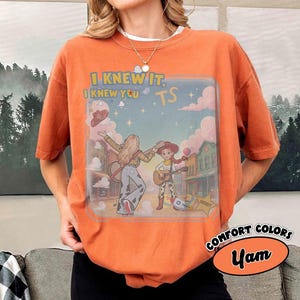 May include: An orange short-sleeve t-shirt featuring a graphic print of two characters playing guitars in a western town setting. The text reads I KNEW IT, I KNEW YOU TS. The design includes clouds, stars, and a sheriff star badge icon.