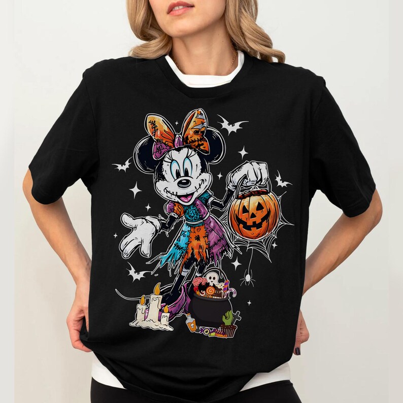 May include: Black t-shirt featuring a Halloween-themed Minnie Mouse graphic. Minnie is dressed in a patchwork costume and holding a jack-o'-lantern. Bats, stars, and candles complete the spooky design.
