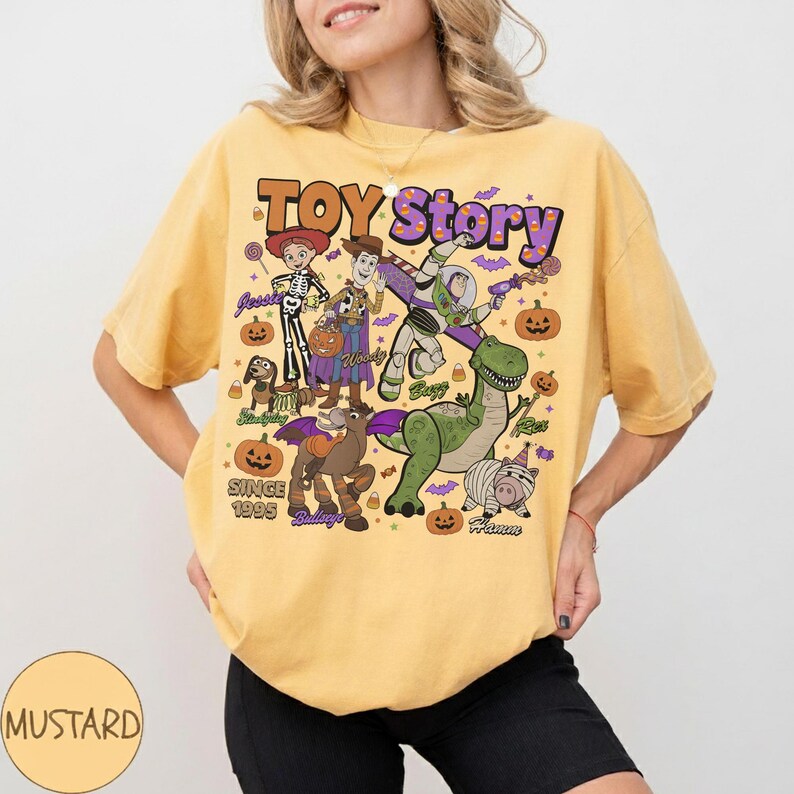Op de afbeelding: Een mosterdgeel t-shirt met een grafische print van Toy Story-personages in Halloween-kostuums. Het ontwerp bevat Jessie, Woody, Buzz Lightyear, Slinky Dog, Bullseye, Rex en Hamm, met pompoenen, vleermuizen en de tekst Toy Story Since 1995.