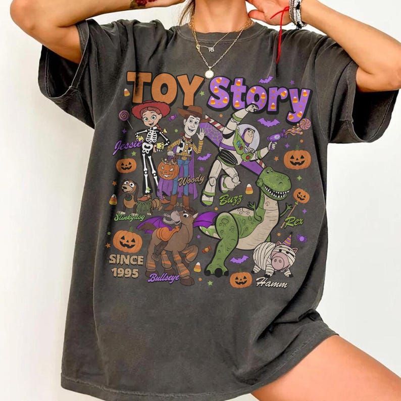 Op de afbeelding: Een antracietgrijs T-shirt met korte mouwen en een Toy Story-afbeelding in Halloween-thema. De print bevat de personages Jessie, Woody, Buzz Lightyear, Rex, Slinky Dog, Bullseye en Hamm, omringd door pompoenen, maissnoepjes en de tekst TOY Story SINCE 1995.