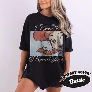 May include: A black short-sleeve t-shirt featuring a rectangular graphic print of a cartoon character with red hair in braids. The text "EST 2026", "I Knew It", and "I Knew You" is printed in a white script font above and below the central image.