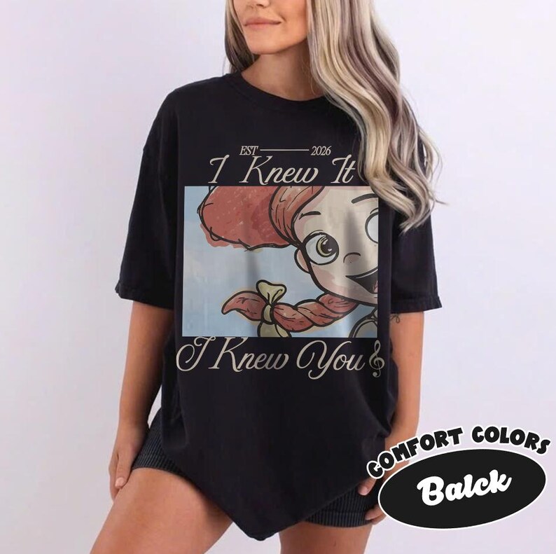 May include: A black short-sleeve t-shirt featuring a rectangular graphic print of a cartoon character with red hair in braids. The text "EST 2026", "I Knew It", and "I Knew You" is printed in a white script font above and below the central image.