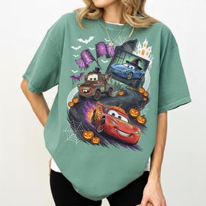 K&ouml;nnte beinhalten: Ein salbeigr&uuml;nes Kurzarm-T-Shirt mit einem Halloween-Motiv, das Charaktere aus dem Film Cars zeigt. Das Design umfasst einen braunen Abschleppwagen, ein rotes Rennauto und ein blaues Auto, umgeben von K&uuml;rbissen, Spinnenweben und Flederm&auml;usen.