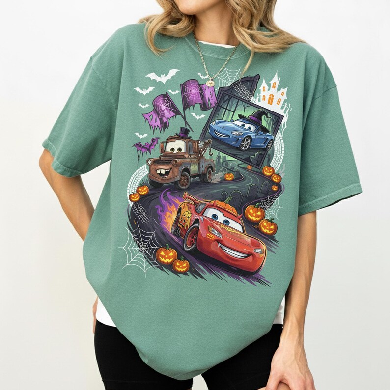 K&ouml;nnte beinhalten: Ein salbeigr&uuml;nes Kurzarm-T-Shirt mit einem Halloween-Motiv, das Charaktere aus dem Film Cars zeigt. Das Design umfasst einen braunen Abschleppwagen, ein rotes Rennauto und ein blaues Auto, umgeben von K&uuml;rbissen, Spinnenweben und Flederm&auml;usen.
