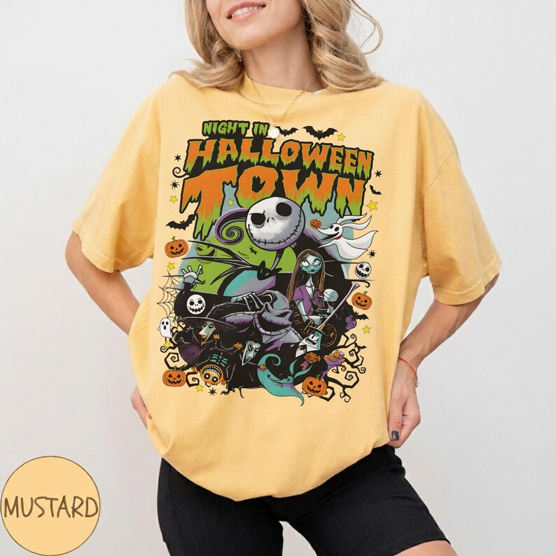 May include: Mustard-colored t-shirt featuring a graphic from "The Nightmare Before Christmas." The design includes Jack Skellington, Sally, and other characters with the text "Night in Halloween Town." The shirt has a crew neck and short sleeves.