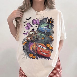 K&ouml;nnte beinhalten: Ein cremefarbenes T-Shirt mit einem Grafikdruck von Charakteren aus dem Film Cars in einer Halloween-Umgebung. Das Design zeigt ein rotes Rennauto, einen braunen Abschleppwagen, ein blaues Auto mit Hexenhut, K&uuml;rbisse, Spinnenweben und Flederm&auml;use.