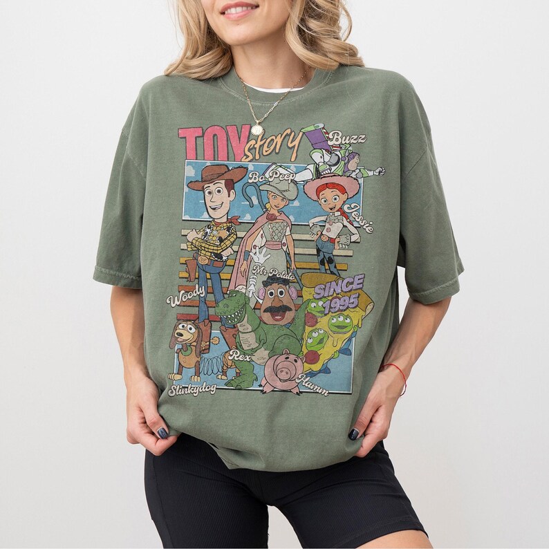 Pu&ograve; includere: T-shirt verde oliva con una grafica dei personaggi del film d'animazione "Toy Story". Il design include Woody, Buzz Lightyear, Jessie e altri personaggi, con il testo "Toy Story" e "Since 1995."